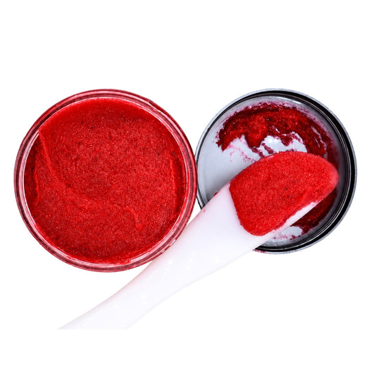 A close-up of a bright red cosmetic or makeup powder in an open round jar, with a white spatula holding a scoop of the powder and a black-lidded container beside it, on a plain white background.