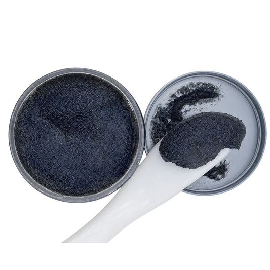 A close-up of a black textured scrub or balm inside an open round metal tin, with a white spatula holding a dollop of the product on a plain white background.