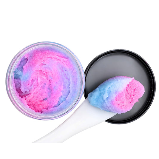 A round clear jar filled with pink and blue swirled cosmetic scrub or shaving cream, shown open with a black lid and a white spatula holding a scoop of the colorful product on a white background.