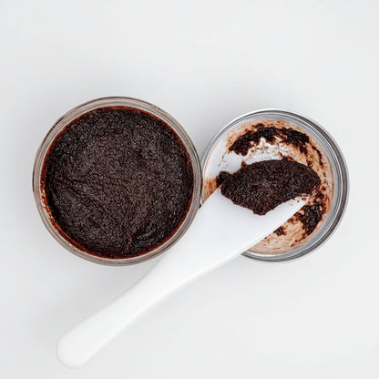 A close-up of a dark brown scrub or cosmetic paste shown in an open round jar with a white spatula, on a plain white background.