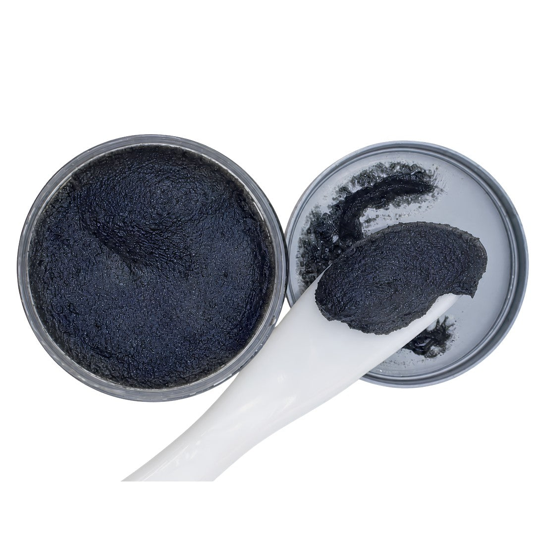 A close-up of a black textured scrub or balm inside an open round metal tin, with a white spatula holding a dollop of the product on a plain white background.