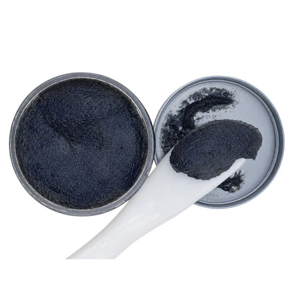 A close-up of a black textured scrub or balm inside an open round metal tin, with a white spatula holding a dollop of the product on a plain white background.