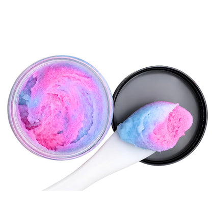 A round clear jar filled with pink and blue swirled cosmetic scrub or shaving cream, shown open with a black lid and a white spatula holding a scoop of the colorful product on a white background.
