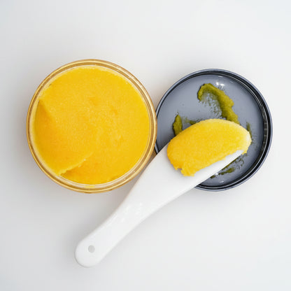 A close-up of a bright yellow cream or balm in an open round container, with a white spoon holding a scoop of the product and a dark lid resting beside it on a white background.