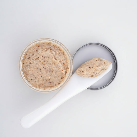 A round clear jar filled with a beige textured scrub or balm, shown next to a white spatula holding a dollop of the product and a silver metal lid on a white background.